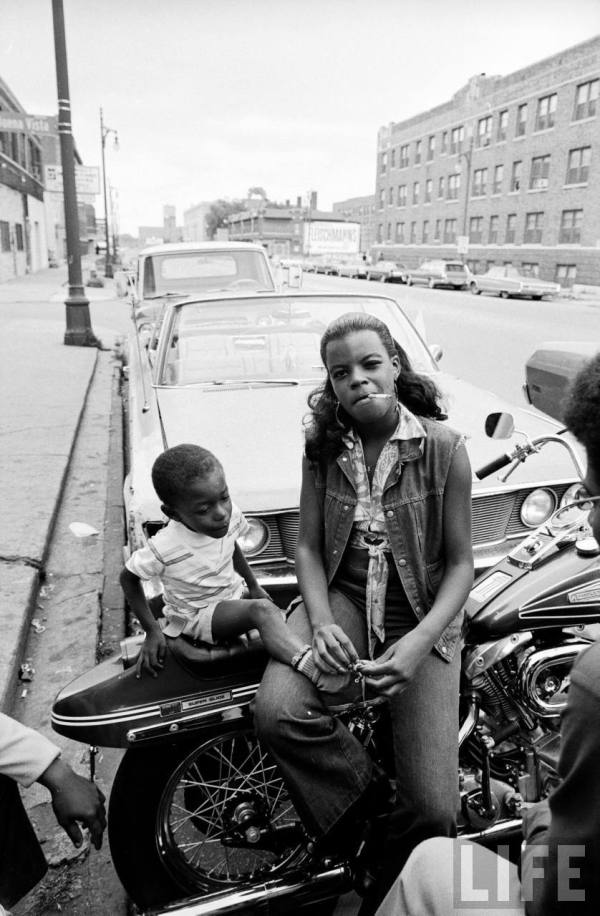 African American woman sitting on a motorcycle w. her child, part of the growing numbers of black motorcycle enthusiasts-- circa 1971.